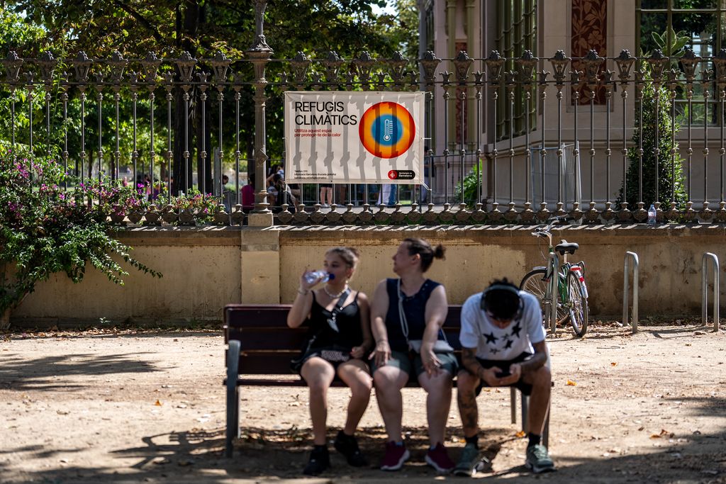 Tres persones assegudes a l'ombra d'un banc. De fons, la cartelleria informativa a un refugi climàtic